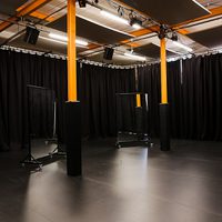 Empty black-box studio space with black curtains, orange support beams, overhead lighting panels, and two rolling transparent screens on a dark floor.