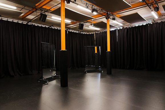 Empty black-box studio space with black curtains, orange support beams, overhead lighting panels, and two rolling transparent screens on a dark floor.