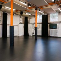 Empty black-box studio space with black curtains, orange support beams, overhead lighting panels, and two rolling transparent screens on a dark floor.