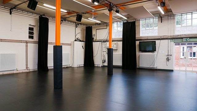 Empty black-box studio space with black curtains, orange support beams, overhead lighting panels, and two rolling transparent screens on a dark floor.