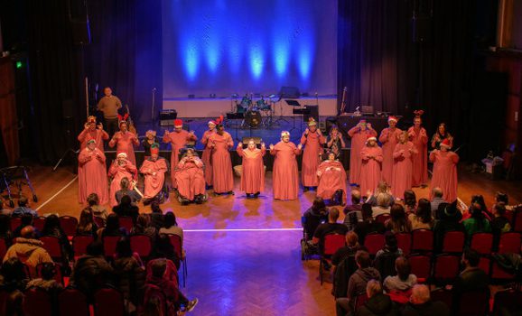 A hall without a stage has performers wearing long red capes performing something. The lights are projecting blue light and rows of people sit on chairs, clear space going down the middle creating a catwalk effect.