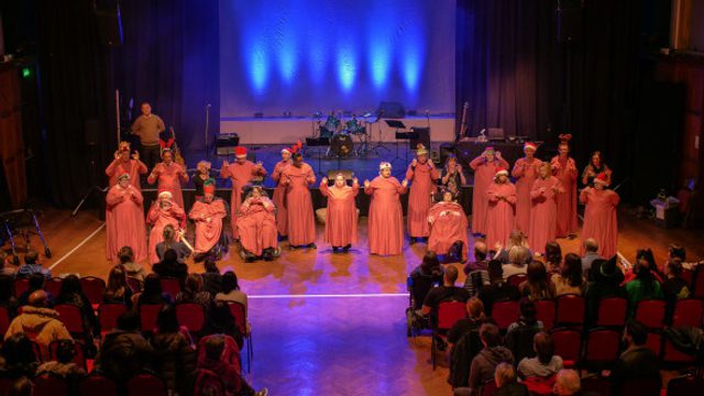 A hall without a stage has performers wearing long red capes performing something. The lights are projecting blue light and rows of people sit on chairs, clear space going down the middle creating a catwalk effect.