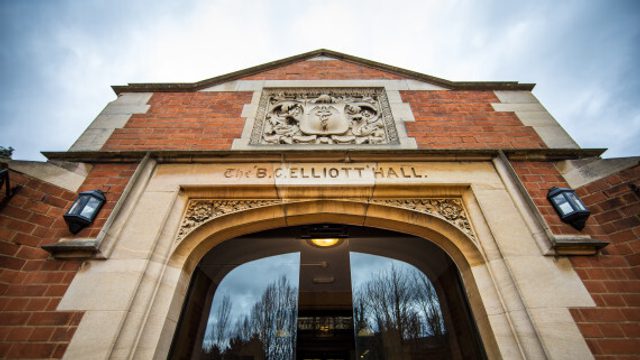Picture of the entrance to the Elliott Hall. It has modern sliding doors that open when you approach it, dark red brick and words carved above the door reading 'THE ELLIOTT HALL'