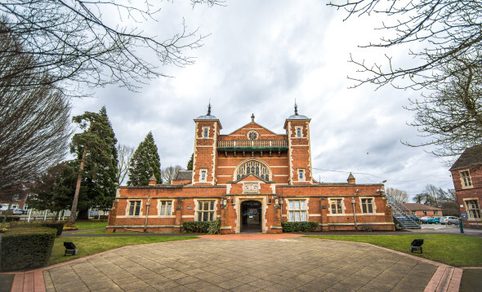 Front view of a red-brick historic building with towers and an arched central entrance, set behind a paved courtyard with trees and lawns under an overcast sky.