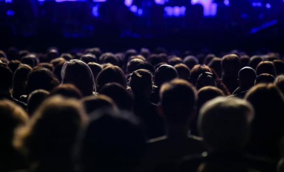 An audience is sat down showing the back of their heads in a dark lit auditorium.