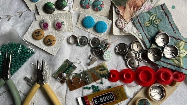 On a white table, there are a set of greena nd yellow pliers, along with patterned buttons, bottle tops and beads ready for jewellery making