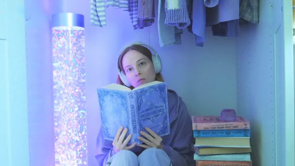 A woman wearing blue jeans and a purple jumpe sits in a lilac painted closet next to a large lava lamp and a pile of books. Some clothes hang up above her head. She is reading a book and wearing overhead headphones