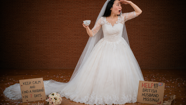 A lady in a white wedding dressing stands, holding a tea cup and gazing into the distance. She stands behind plaquards that say 'keep calm and find a husband in 7 days' and 'Help! British husband missing!'