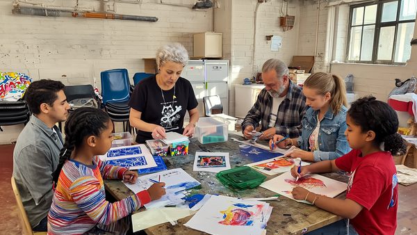 A small group of children and two adults sit around a large table in a workshop space, working on art projects. They are drawing, coloring, and cutting paper, with art supplies like markers, scissors, and printed images spread across the table. The instructor stands at the center, guiding the activity, while participants focus on their individual creations in a bright, casual studio setting.