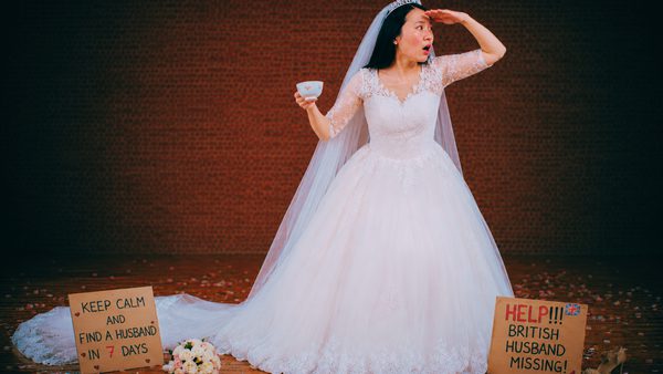 A lady in a white wedding dressing stands, holding a tea cup and gazing into the distance. She stands behind plaquards that say 'keep calm and find a husband in 7 days' and 'Help! British husband missing!'