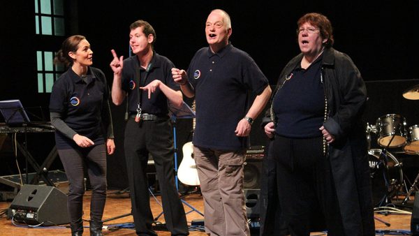 From left to right: a teacher, two men and a woman, all wearing black, are seen on stage singing and dancing.