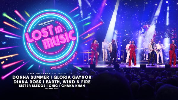 The title in bright pink neon letters 'Lost in Music' is shown on the left inside a circle of colourful blue, pink and green neon lights. An image of singers on stage with a large audience is shown on the right. The singers are wearing red jumpsuits and are bathed in theatrical lighting.