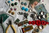 On a white table, there are a set of greena nd yellow pliers, along with patterned buttons, bottle tops and beads ready for jewellery making