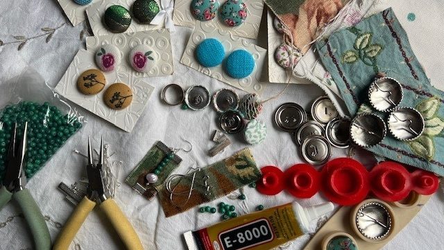 On a white table, there are a set of greena nd yellow pliers, along with patterned buttons, bottle tops and beads ready for jewellery making