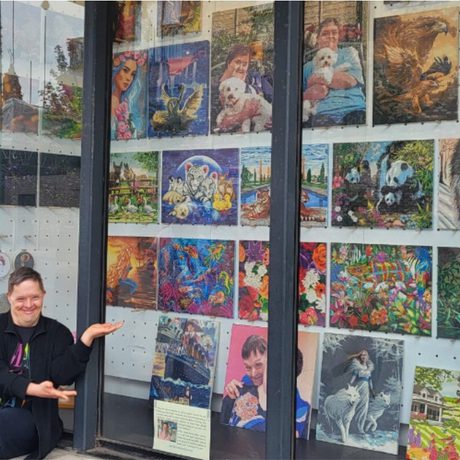 Person sitting outside a glass display window filled with colorful paintings, smiling and gesturing toward the artwork. The display includes portraits, animals, fantasy scenes, and nature illustrations arranged in neat rows inside the gallery window.