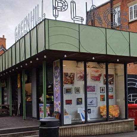 Small green kiosk-style building with large glass windows displaying artwork, set in a modern urban plaza.