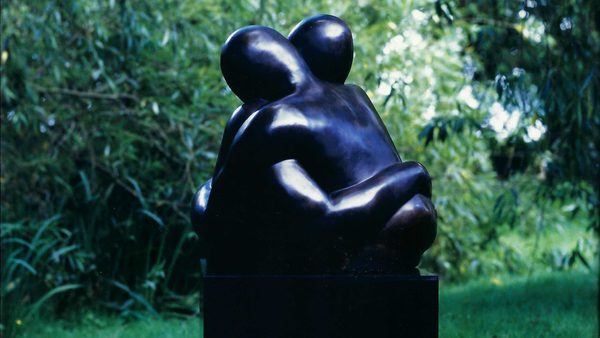 A sculpture of two figures made from black stone in an embrace. They are displayed outdoors surrounded by greenery.