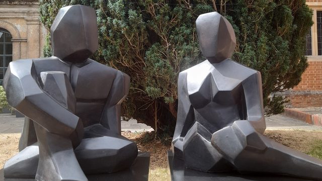 A sculpture of 2 geometric figures made from dark grey stone materials. The sculpture sits in front of a large tree.