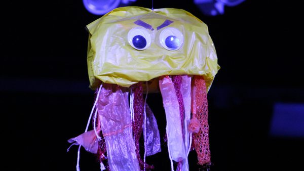 A puppet jellyfish made entirely out of recycled plastic. It has a yellow top and purple tentacles, with googly eyes and an angry expression on it's face!