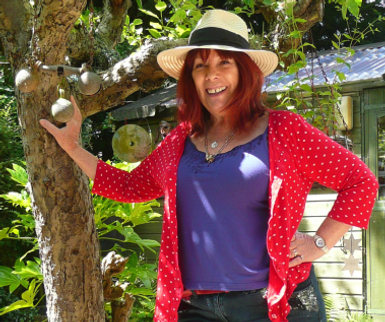 A woman poses in front of tree wearing a sun hat, purple top and red cardigan with white polka dots.