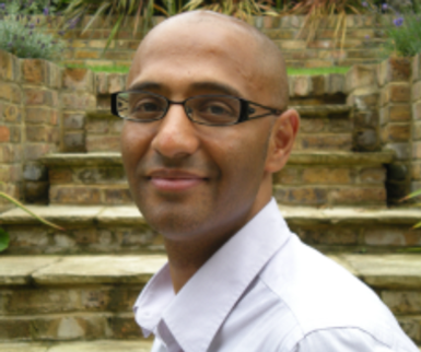 Bald man with glasses closed mouth smiles wearing a white collared shirt.