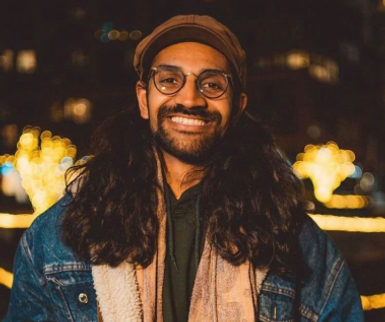 Smiling person with long dark hair wearing round glasses and a brown cap, dressed in a denim jacket layered over a hoodie and scarf, standing outdoors at night with warm, blurred lights in the background.