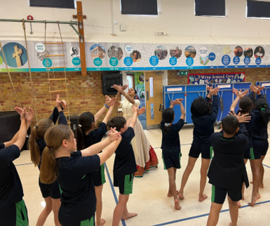 A group of children in sports uniforms stand in a hall, following a dance instructor positioned in front of them. Their arms are raised and angled outward in matching poses. The background shows gym equipment, a brick wall, posters, and blue folding screens.