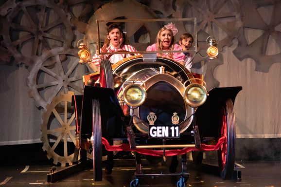 Three performers on a theatre stage sit in a vintage-style car prop, looking shocked and excited as if speeding forward.