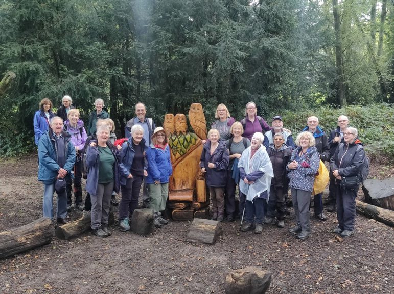 A group of elderly people pose next to a wooden sculpture surrounded by nature
