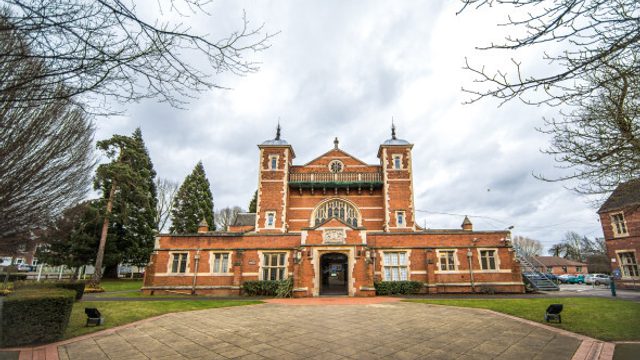 Front view of a red-brick historic building with towers and an arched central entrance, set behind a paved courtyard with trees and lawns under an overcast sky.