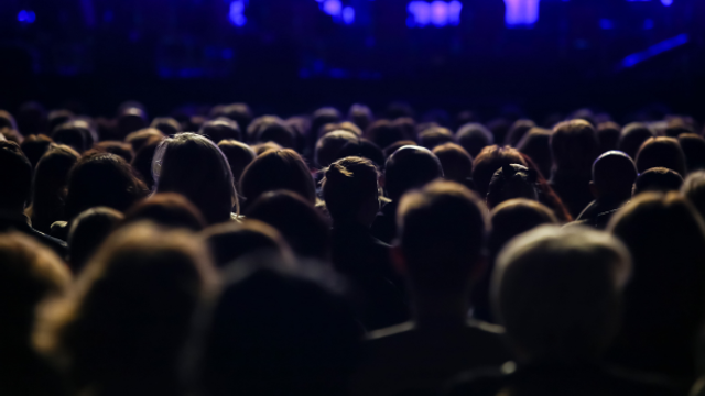 An audience is sat down showing the back of their heads in a dark lit auditorium.