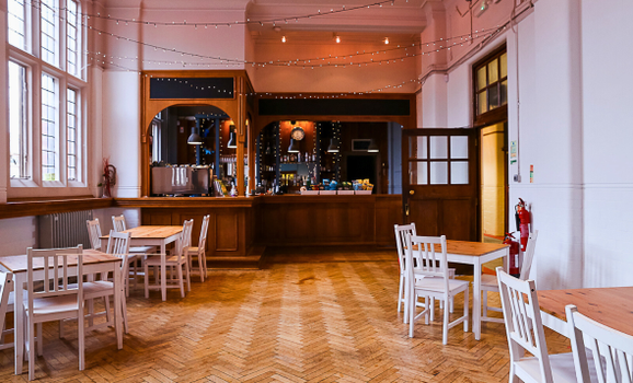 A spacious café interior with high white walls and large windows, featuring a polished herringbone wooden floor. Several small wooden tables with white chairs are arranged around the room. In the background, a wooden bar counter with shelves and equipment is softly lit, and string lights are draped across the ceiling, creating a warm, cosy atmosphere.