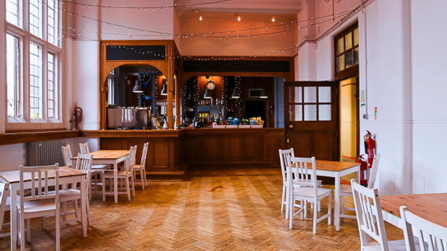 A spacious café interior with high white walls and large windows, featuring a polished herringbone wooden floor. Several small wooden tables with white chairs are arranged around the room. In the background, a wooden bar counter with shelves and equipment is softly lit, and string lights are draped across the ceiling, creating a warm, cosy atmosphere.