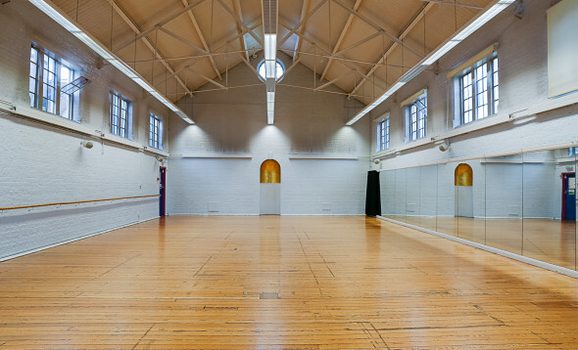 A large, empty dance or fitness studio with a polished wooden floor and high vaulted ceiling with exposed beams. White brick walls line the space, with tall rectangular windows on both sides letting in natural light. One wall is covered with floor-to-ceiling mirrors and a wooden ballet barre, and fluorescent strip lights run along the ceiling.