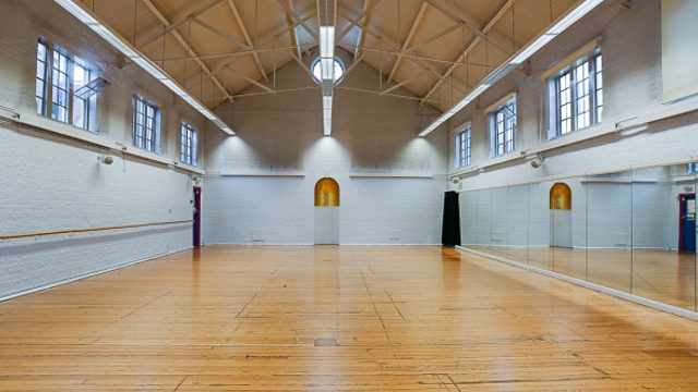 A large, empty dance or fitness studio with a polished wooden floor and high vaulted ceiling with exposed beams. White brick walls line the space, with tall rectangular windows on both sides letting in natural light. One wall is covered with floor-to-ceiling mirrors and a wooden ballet barre, and fluorescent strip lights run along the ceiling.
