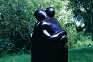 A sculpture of two figures made from black stone in an embrace. They are displayed outdoors surrounded by greenery.