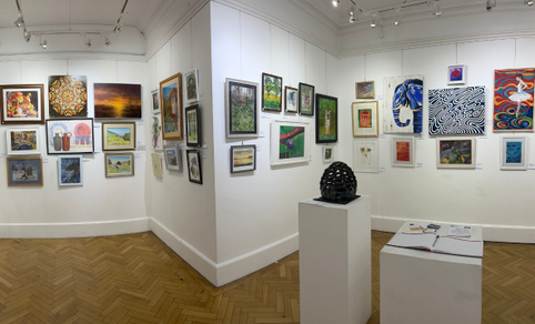 Interior of a white-walled art gallery showing a corner view with many framed paintings hung closely together, two small sculptures on pedestals, and a wooden herringbone floor under track lighting.
