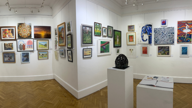 Interior of a white-walled art gallery showing a corner view with many framed paintings hung closely together, two small sculptures on pedestals, and a wooden herringbone floor under track lighting.