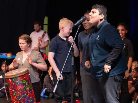 Kids peform on stage with a microphone as a person plays African drums in the background.