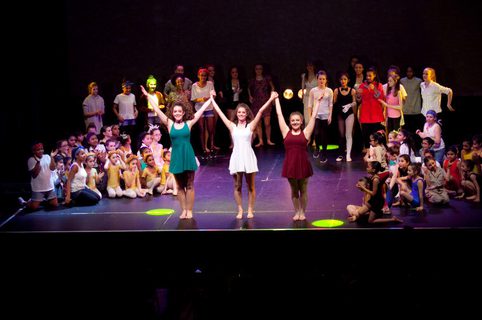 3 performers take a bow on stage in front of an audience of people and performers on edges of stage.