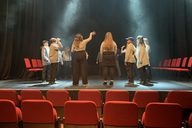 Young people on stage in an empty auditorium stand in a circle while lights and haze shine down on them.
