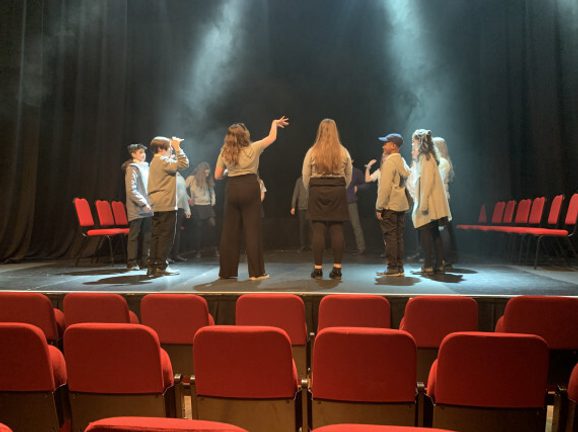 Young people on stage in an empty auditorium stand in a circle while lights and haze shine down on them.