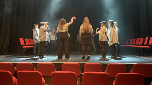 Young people on stage in an empty auditorium stand in a circle while lights and haze shine down on them.