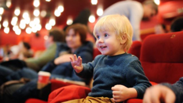 Little boy in sat in a theatre smiling and waving at something he is looking at taking place on stage