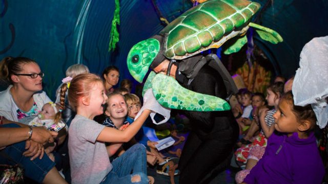 Kids laugh and smile looking at a puppet of a turtle.