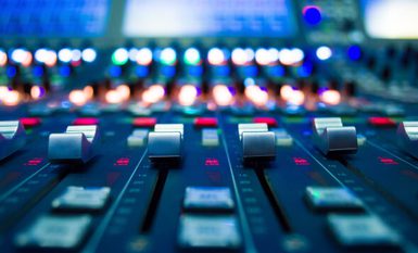 Close-up of an audio mixing console with multiple faders and glowing LED indicators, shot with shallow depth of field and blue-toned lighting.