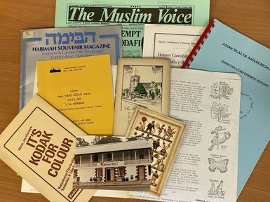 A selection of archival publications and community materials spread across a table, including The Muslim Voice newspaper, a Habimah souvenir magazine from Stanmore & Canons Park Synagogue, a report on Asian health awareness, and a Gujarati-language booklet from the London Borough of Harrow. Also visible are a Kodak photography booklet, an illustrated nature guide page, and a historic photograph of a building.