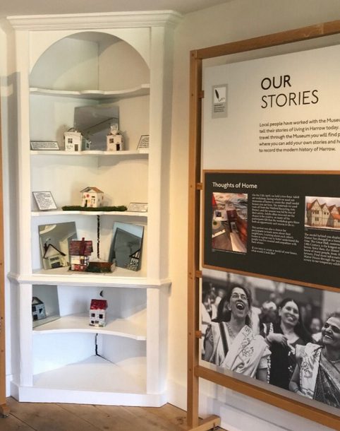 A museum display showing small model houses arranged on white corner shelves beside an interpretation panel titled “Our Stories.” The panel includes text about local people sharing their experiences of living in Harrow, with photographs and information about community stories.