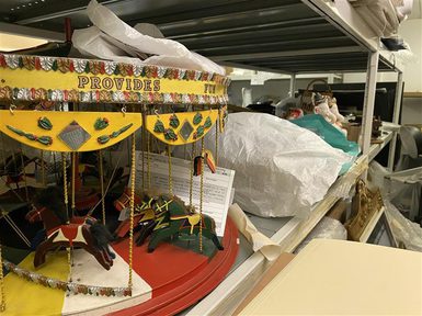 A brightly painted model carousel with small painted horses hanging from poles, decorated with yellow panels and the words “PROVIDES FUN” around the canopy. The carousel sits on a shelf inside a museum store, surrounded by wrapped and boxed objects and metal storage shelving.