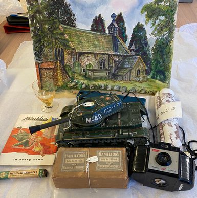 A selection of museum objects arranged on a table, including a green toy tank marked “M-40,” a Kodak Brownie 127 camera, two boxes of Hamilton’s varnish brushes, a small wooden pipe in a clear bag, a medicine dropper box, a printed Aladdin booklet, a rolled textile, and a small glass. Behind the objects is a framed painting of a church set in a wooded landscape.